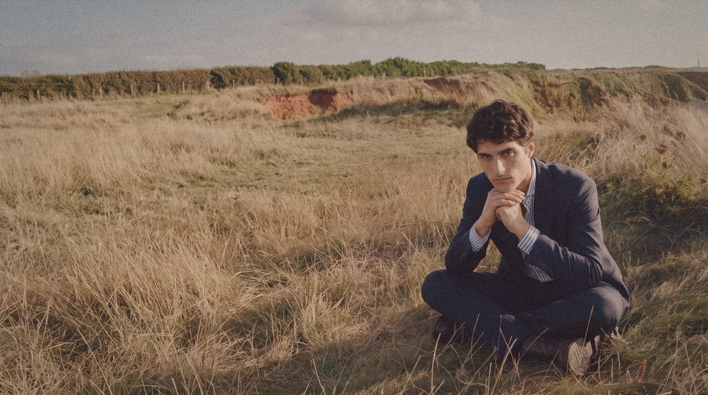 A man in a navy suit sits cross legged in tall dry grass.